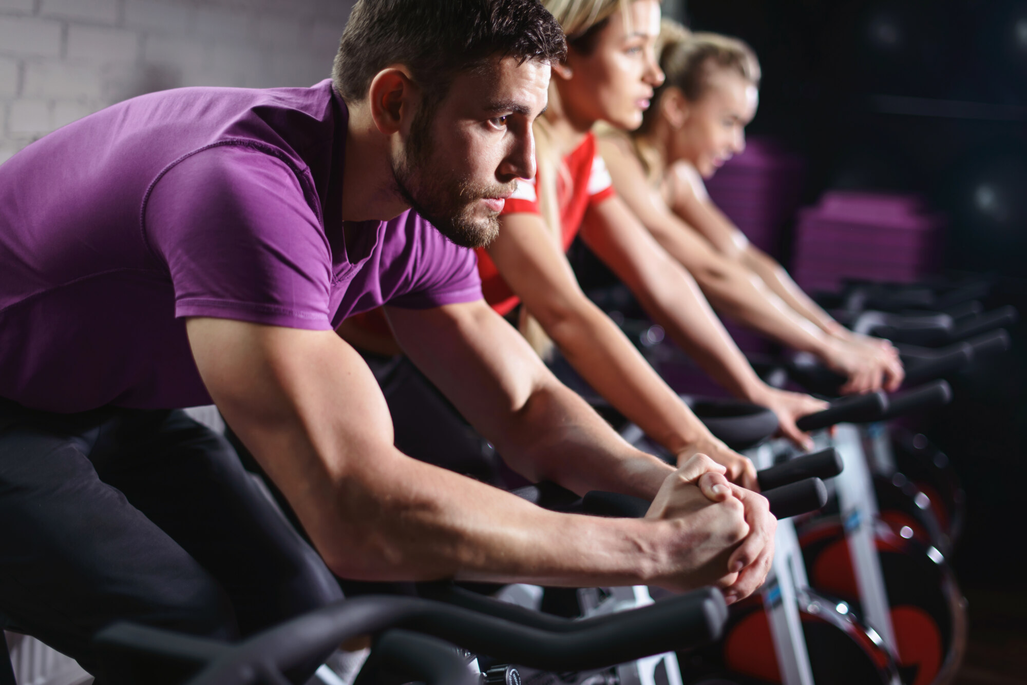 Close up hands of man biking in spinning class. Group of smiling friends at gym exercising on stationary bike. Happy cheerful athletes training on exercise bike.